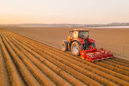 PRAGUE , CZECH REPUBLIC - MARCH 18 2022: Heavy tractor uses plow to make beds for planting potatoes in agricultural field. Powerful machine operates at farm on sunny day aerial viewのeditorial素材