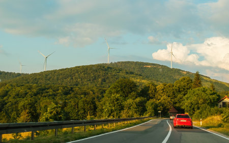 Red car drives on road against green hillside with windmill farm. Producing ecological electrical energy among green landscape under blue skyの写真素材