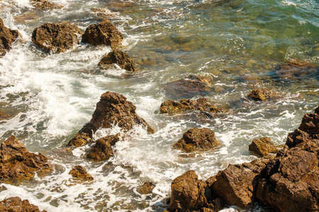 Small ocean waves crashing of rocks on shallow coastline at high tide at bright sunlight. Transparent waves create foams rolling on stonesの写真素材