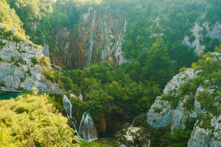 Waterfalls with cascades falling from steep rocks into canyon. Forestry and bare cliffs surround gorge with clear blue lake on bottom of Plitvice lakes upper viewの写真素材