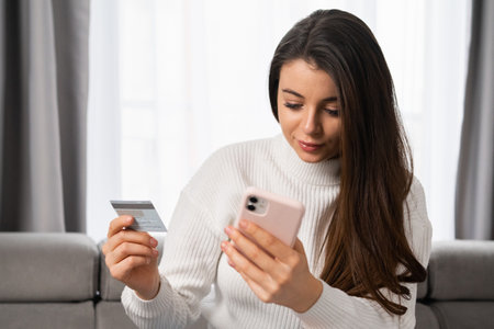 A caucasian woman with long dark hair pays by card using her mobile phone at home with a modern...