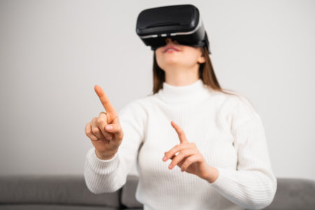 Curious housewife in VR goggles swipes virtual interface to choose game. Young woman rests on couch tasting modern innovations at homeの写真素材