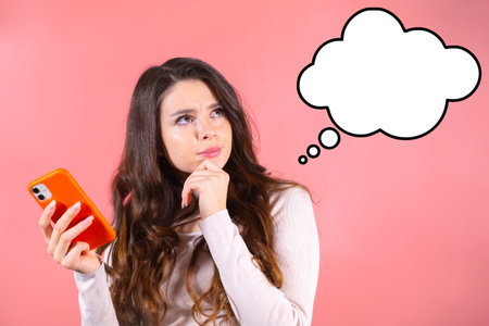Thoughtful girl with a phone thinks about shopping and a cloud for text on a pink background