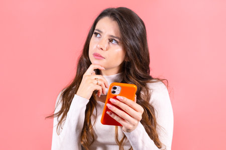 Young woman with amused and smiling expression enjoys scrolling social media on orange smartphone. Long-haired brunette lady smiles looking at feedsの写真素材