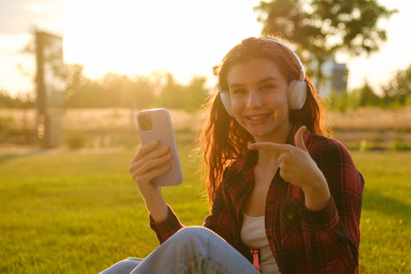 Smiling happy woman in headphones pointed by finger at the smartphone sitting on the grass in the...