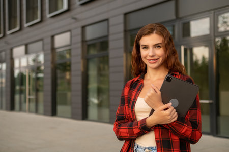 Happy red haired student, wearing casual clothes, holding a tablet, and standing near the university.の写真素材