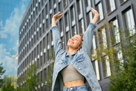 A contented young woman with flowing, loose hair beams with a wide smile, raising her arms in the air. She stretches happily after a day of work beside an office building.の写真素材