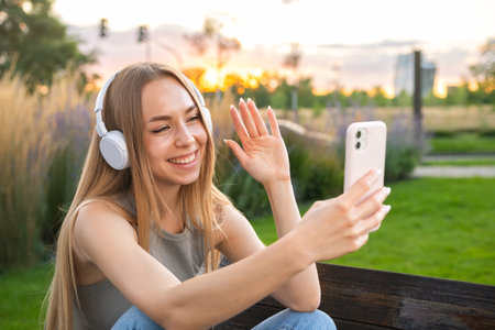 Joyful young woman in headphones talks to friend on video call sitting on park bench in evening. Lady chats with boyfriend on phone in parkの写真素材
