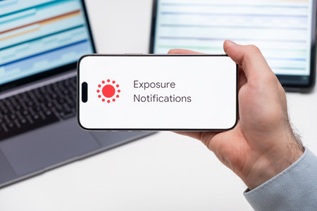 Exposure Notifications application logo on the screen of smart phone in mans hand, laptop and tablet are on the table in the background, December 2023, Prague, Czech Republic.のeditorial素材