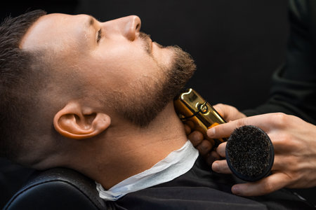 Hairdresser shaves hair on client neck creating beard shape with machine in barbershop closeup. Barber uses electrical shaver in salonの写真素材