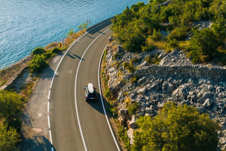 A sporty car with a SUP on the roof speeds along a coastal road, with a rocky hillside on one side and cliffs overlooking the seaの写真素材