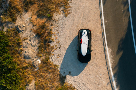 Aerial shot of a compact black hatchback featuring a paddleboard mounted on its roof racks, parked alongside a rustic countrysideの写真素材