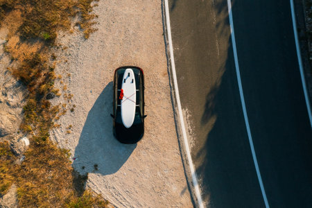Black car with a paddleboard securely fastened on its roof, parked neatly on the shoulder of a winding coastal road.の写真素材