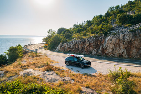 Black car with a SUP lashed onto its roof, stands on a side of a rugged coastline road.の写真素材