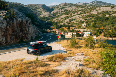 Black car with a paddleboard securely fastened on its roof, parked neatly on the shoulder of a winding coastal road.の写真素材