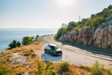 Black car with a paddleboard tightly secured atop its roof, parked in the side of the coastal highway with the shimmering seaの写真素材