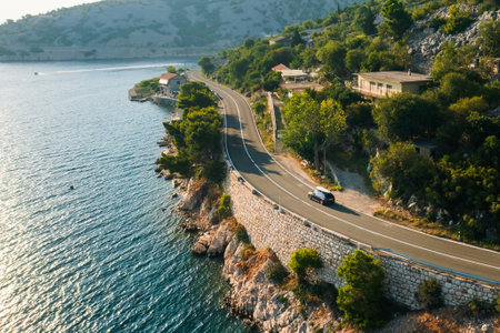 Car with paddleboard on the roof drives along a narrow coastal road under a clear blue sky.の写真素材
