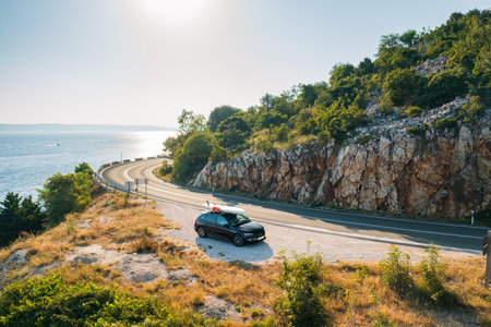 Car with a paddleboard lashed onto its roof, parked on the side of a quiet country road, with the shimmering sea visible through the treesの写真素材