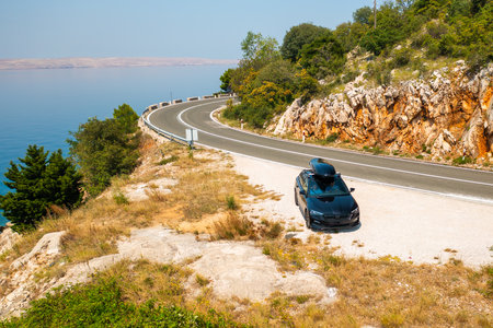 Black car with a roof box, parked at the edge of a narrow, winding road on a rugged cliff.の写真素材