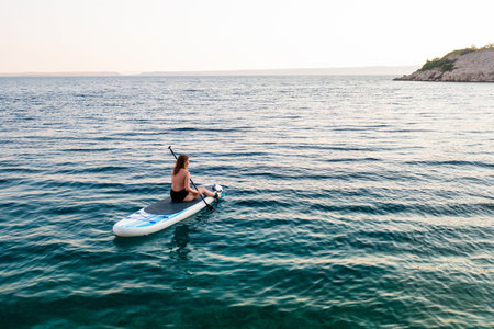 A woman paddle boarding on tranquil waters near a rocky shoreline at sunset.