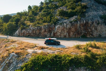 Car with a SUP lashed onto its roof, stands on the side of a country road, near a rugged cliff overlooking the windswept seaの写真素材