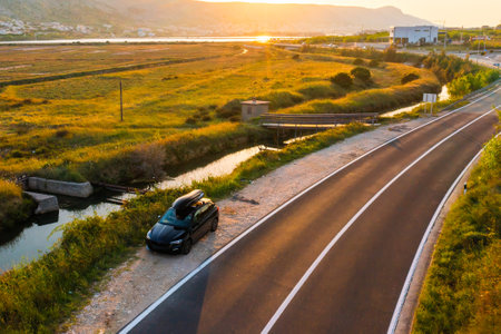 A black car with a roof rack parked alongside a quiet winding road in a rural area during sunset.の写真素材