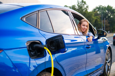 Blonde woman looks from the car window while her electric vehicle charges at a convenient charging point outsideの写真素材