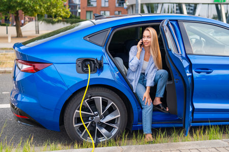 Young woman sits in a blue electric car, charging it while talking on her smartphone.の写真素材