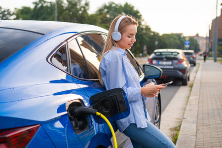 Electric vehicle charging while a blonde woman with long hair, wearing casual clothes, enjoys music with headphones and uses her smartphone in an urban setting.の写真素材