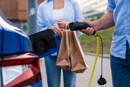 Man plugging in a connector to EV while his wife holds shopping bags at an urban parking lot.の写真素材