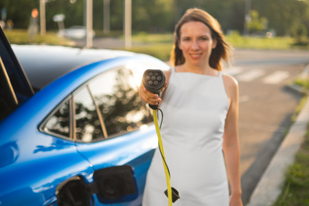 Smiling woman in a white dress holding charger plug in front of her to charge the electric car in the streetの写真素材
