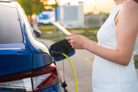 Pregnant woman in white dress charging an electric car and using her mobile phone to payの写真素材