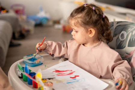 Cute toddler girl painting with gouache at a table near a sunlit window. Natural light highlights her artwork and the joyful atmosphere of creativity.の写真素材