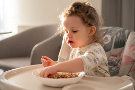 Small curly girl eating cereal at the table in a childs high chair. Natural light and warm home atmosphere.の写真素材