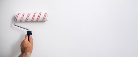 Banner of close up of masculine hand gripping a roller brush, painting a white surface during...