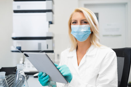 Blonde female scientist wearing a lab mask and rubber gloves recording chemical experiment results...
