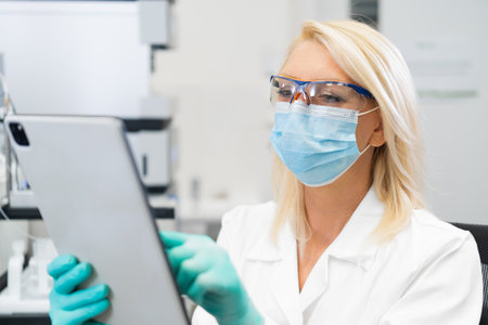 Middle aged female scientist wearing mask and lab coat using tablet in laboratory for research and...