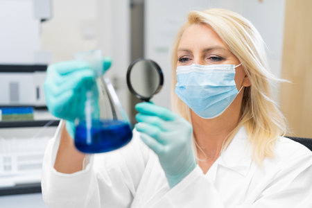 Middle aged female scientist wearing mask in lab, studying chemical solution in flask through magnifying glass.の写真素材