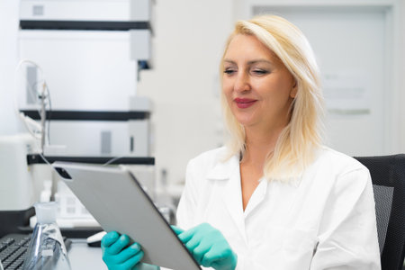 Blonde female scientist without mask and wearing rubber gloves recording experiment results on a tablet in a chemistry laboratory filled with flasks, beakers, and HPLC instrumentsの写真素材
