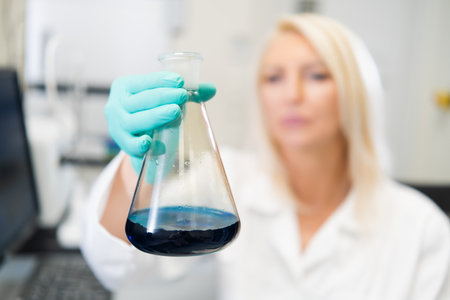 Blonde woman scientist in laboratory wearing lab coat, observing blue liquid in glass flask.の写真素材
