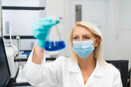 Female scientist wearing mask and lab coat, examining blue chemical solution in glass flask in laboratory.の写真素材