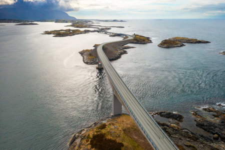 Atlantic ocean road demonstrates impressive span stretching across rocky islets in sea. Highway elegantly curves along path under muted cloudy skyの写真素材