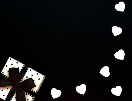 Gift box wrapped with polka dot paper with black ribbon on black background with white hearts. Male holiday concept. Valentineâs Day, Birthday greeting or invitation card. Flat lay with copy space.の写真素材