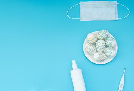 Decorated Easter eggs lie on the plate, medical face mask, thermometer and sanitizer on blue background with copy space. Happy Easter during quarantine, stay home, social distance, COVID-19 concepts.の写真素材