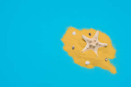 White starfish on bright yellow sand with small sea shells on blue background. Summer time,...