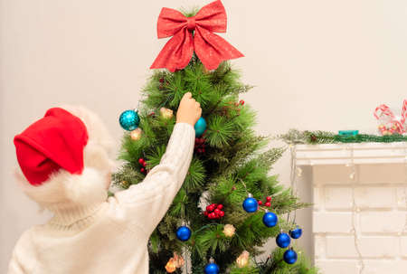 Child boy in red Santa hat decorates Christmas tree close up. Xmas or New Year cozy holidays...