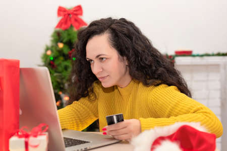 Happy woman holding credit card and shopping online on laptop against Christmas tree in the background. Xmas online shopping, preparing for the New Year, buying present, winter holiday sales conceptの写真素材