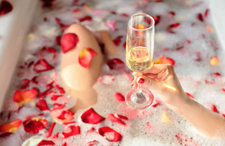 Female hand holding a glass of white wine while sitting in a bathtub with foam and red rose petals....