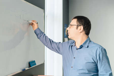 Man employee in blue shirt and glasses writing something on white board during business meeting in the office. Business, busy working day conceptの写真素材