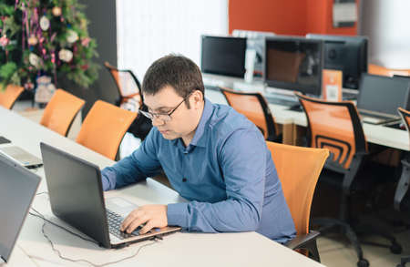 Man employee in blue shirt and glasses sitting at the desk in office using laptop computer....